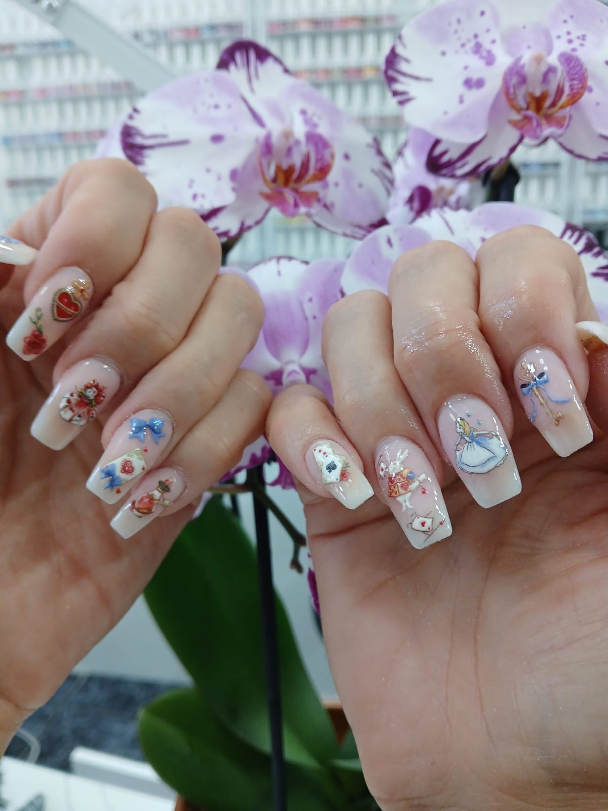Twee handen met lichte nagels vol kleine nail art, met orchideeën op de achtergrond